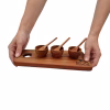 Manos de mujer sosteniendo una tabla de madera de 28x12 cm con tres pocillos y cucharitas, lista para servir en la mesa.