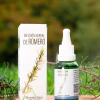 Frasco gotario de vidrio verde y caja blanca de la Infusión Herbal de Romero (Rosmarinus officinalis), sobre una base de madera rodeada de ramas frescas de romero en un entorno natural.