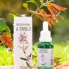 Frasco gotario y caja de la Infusión Herbal de Tomillo (Thymus vulgaris), sobre una base de madera en un entorno natural con flores.