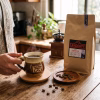 Persona sosteniendo una taza de cerámica rústica con café caliente junto a un empaque de 1 kilo de café Antigua Landorff y un moledor clásico.